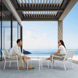 Two people relax on white aluminium patio chairs with one using a matching footrest, all set around a sleek side table with a ceramic glass top. This stylish alfresco furniture set is placed in the outdoor area of a modern luxury home on a sunny day, creating a perfect spot for relaxation.
