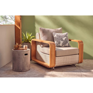 A cozy living room featuring a modern, light grey outdoor wicker lounge chair with wooden arms, positioned near a large window. The room includes a stylish concrete coffee table, potted plants, and warm, inviting decor.