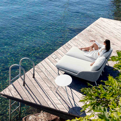 A lady relaxing on a comfortable chaise lounge, set on a wooden deck overlooking a serene river, with a round side table beside her, offering both style and functionality in this peaceful outdoor living space.