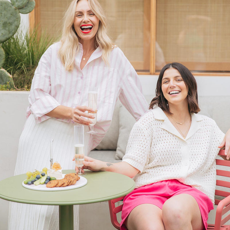 Two women enjoying food and drinks around a green Nardi Spritz round dining table with one pink chair, creating a vibrant alfresco furniture setup in a backyard area.