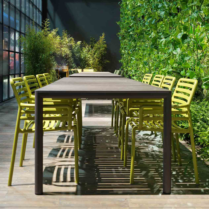 Three black Nardi Tevere outdoor extension dining tables in a modern common area, surrounded by green outdoor chairs and lush greenery, creating a chic alfresco furniture setting.