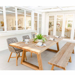 A modern wooden outdoor dining set featuring a rectangular wooden table with a light finish, 3 outdoor wicker chairs on one side, and a large outdoor teak bench on the other side. The furniture is set on a modern patio made of stone.