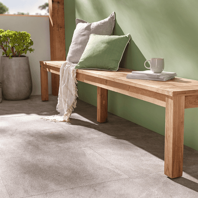 The light-brown 270 cm long wooden 'Polly' outdoor bench (made from recycled teak), one of Remarkable Outdoor Living's best pieces of outdoor furniture, is set against a green wall with the sun shining through. On the bench are some throw pillows, a throw blanket, some books, and a tea cup. In the background, against a window, there is a pot plant as well.