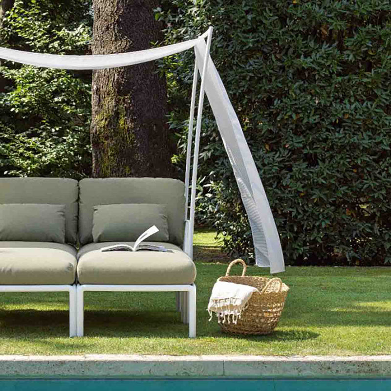 Two-seater outdoor furniture lounge with a white frame, green cushions, and a white flowing fabric canopy, set poolside in a sunny backyard with greenery in the background and a woven basket placed beside it.